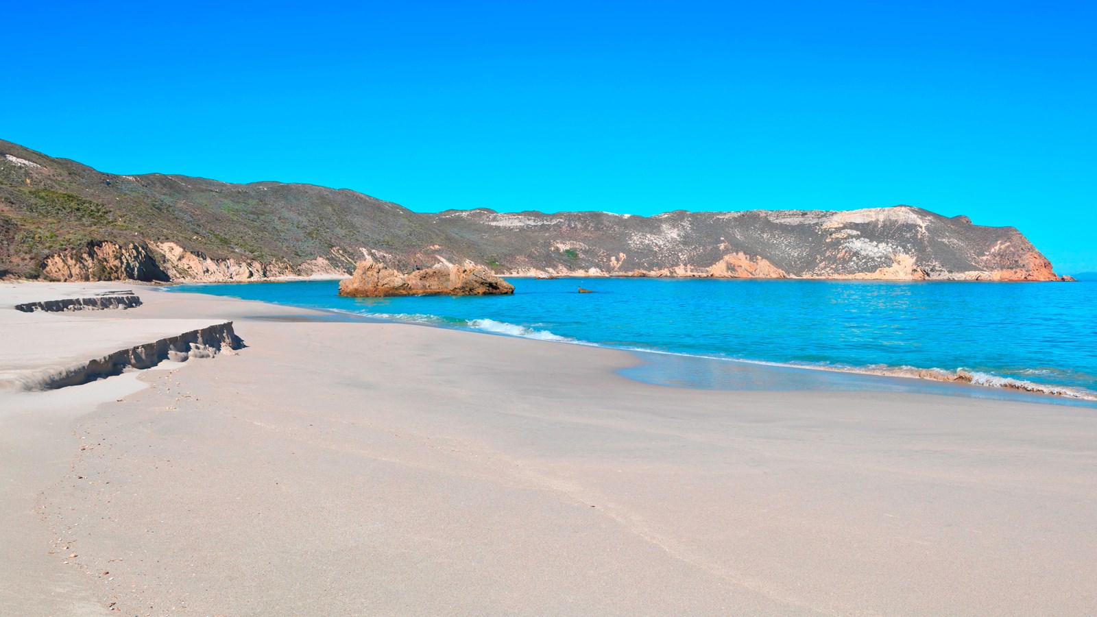 white sand beach in harbor with rock in foreground and cliffs in background.