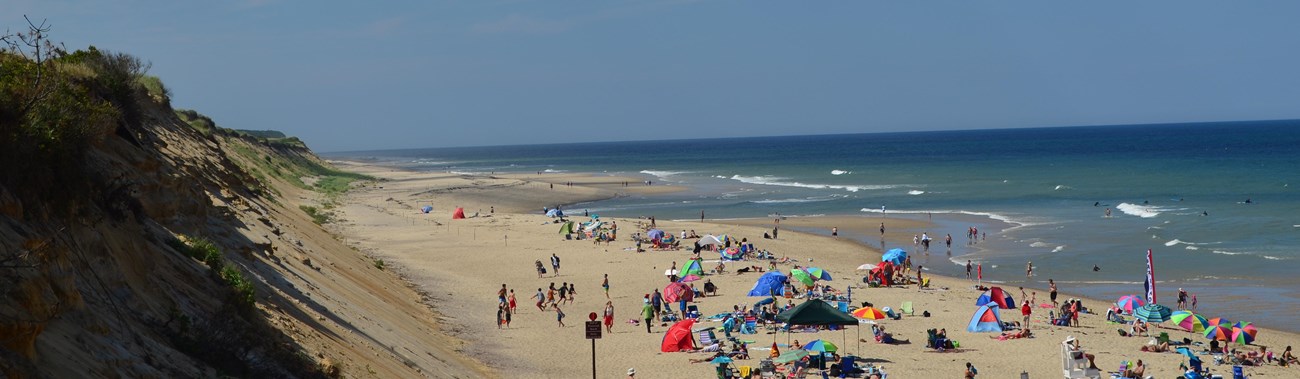 A crowd of people enjoy a wide sandy beach under light clouds and a blue sky.