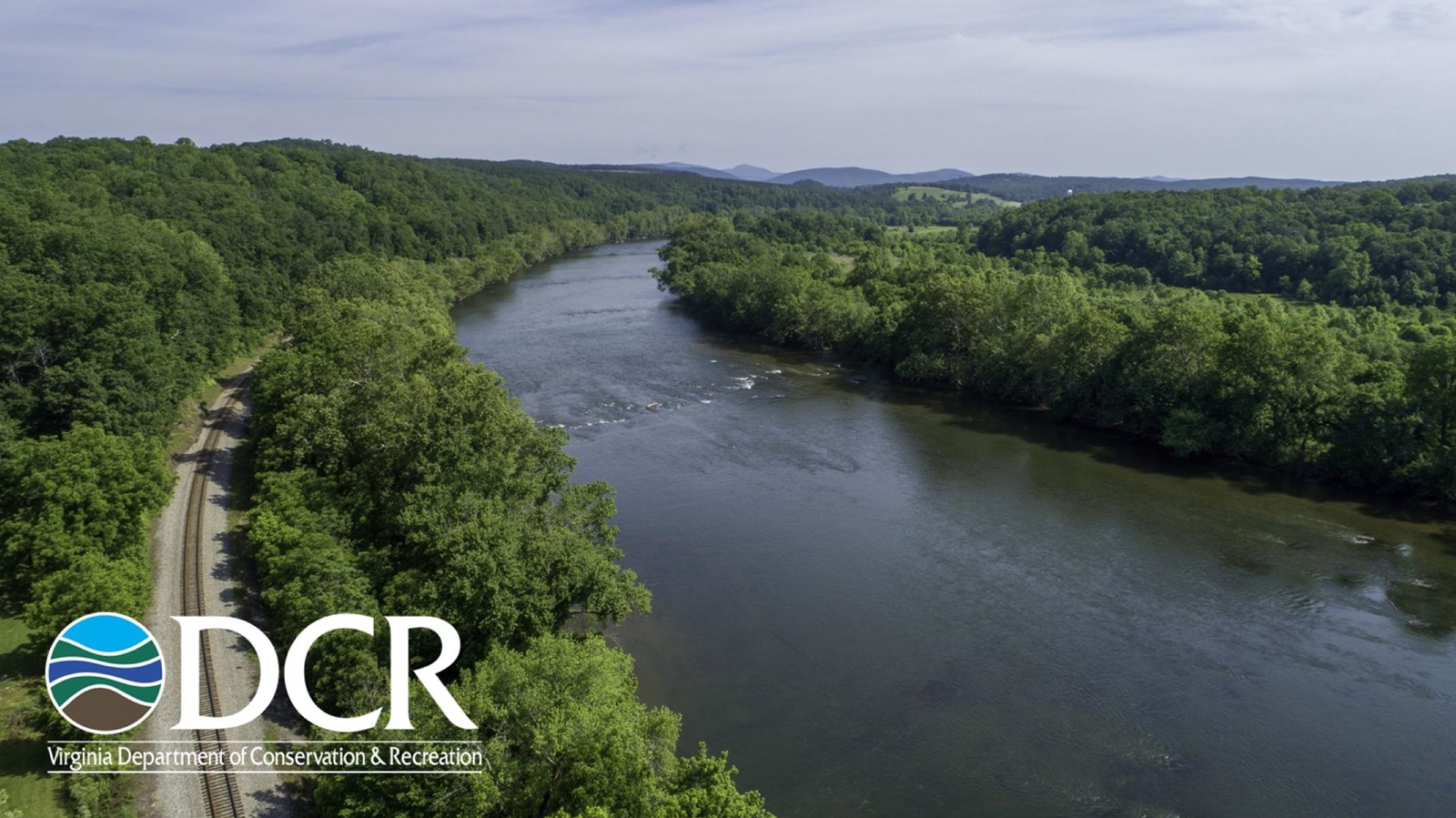 A winding river flows through a green landscape, bordered by trees and hills, with a railway track.