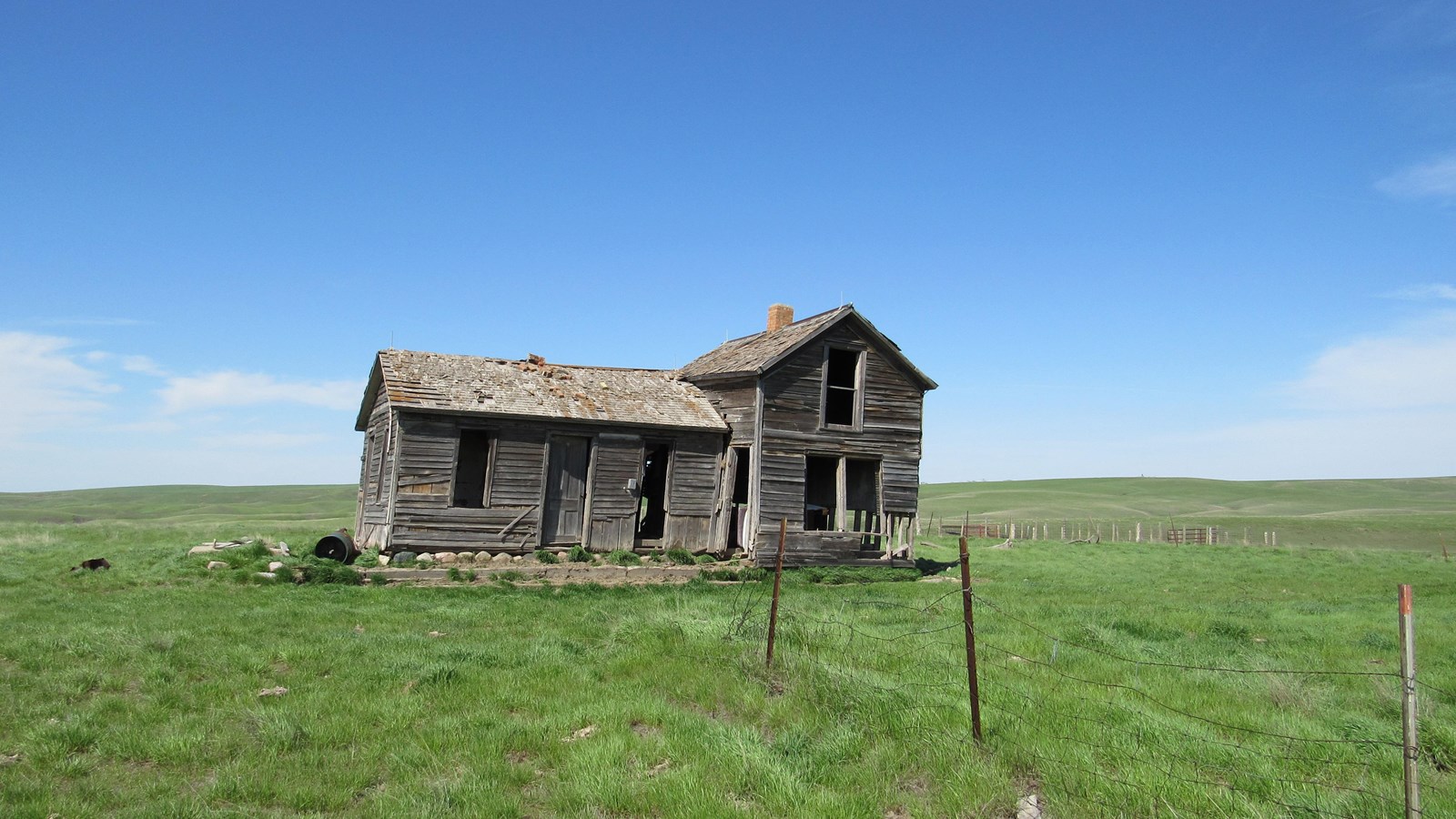 An abandoned weathered wood-frame home sits in a green flat meadow that stretches to the horizon.