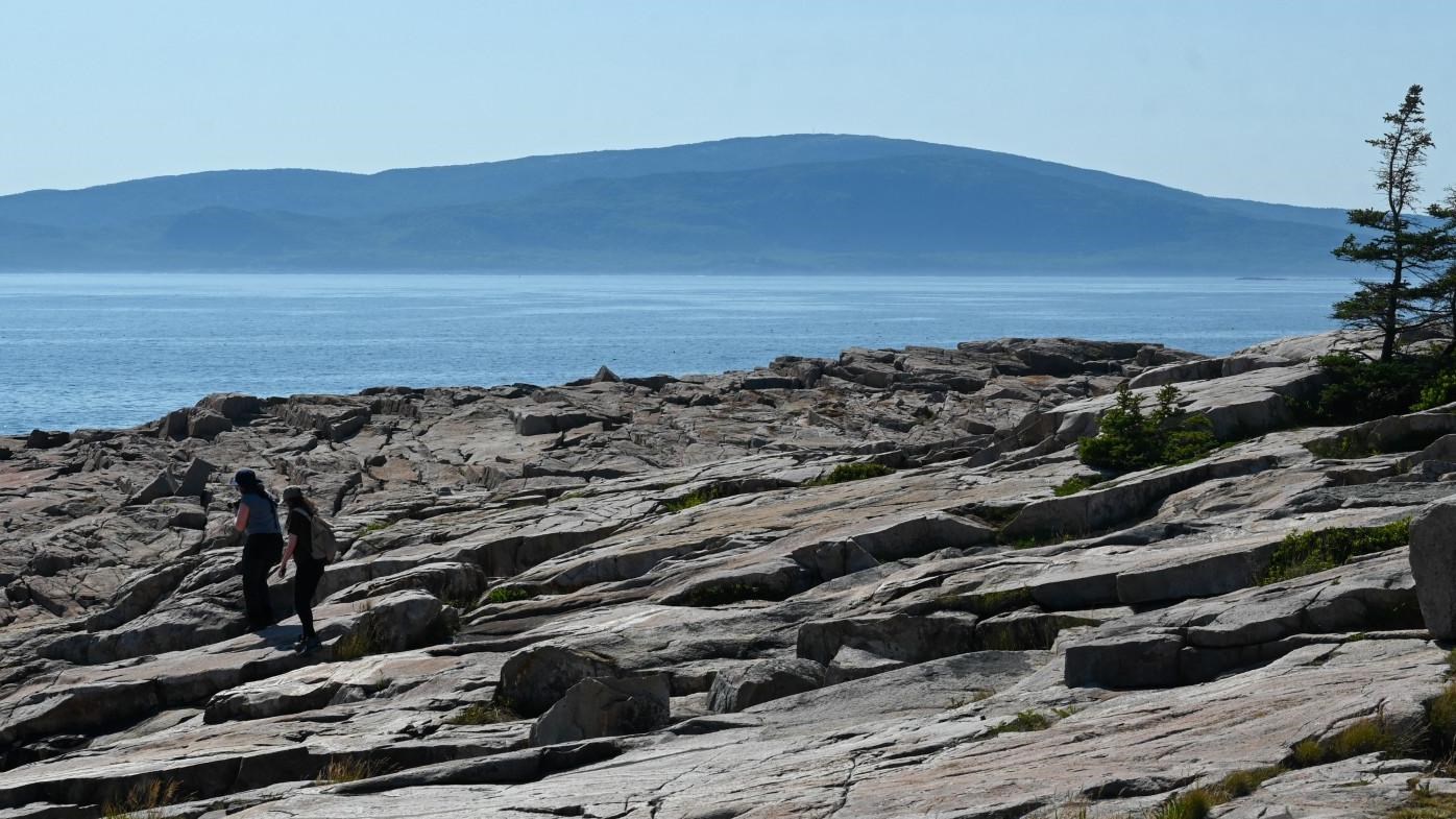 Two women walking on a rocky shore with water and a mountain the backgound