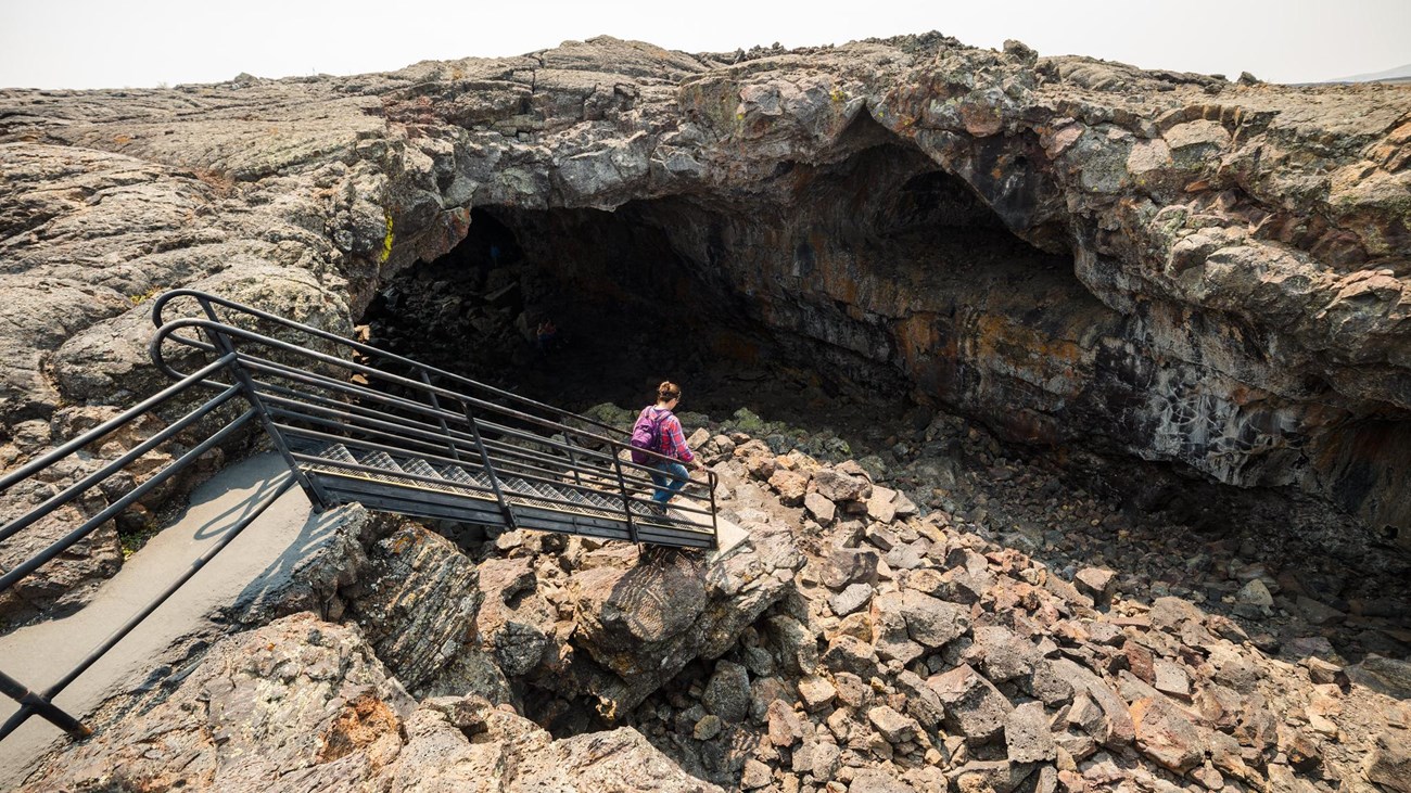 A person walks down a set out stairs into the wide open entrance of a lava tube.