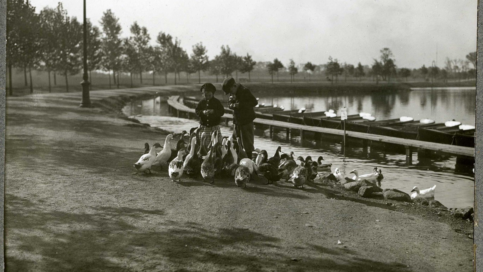 Black and white of lake with two children on edge feeding group of ducks. Canoes on edge of lake