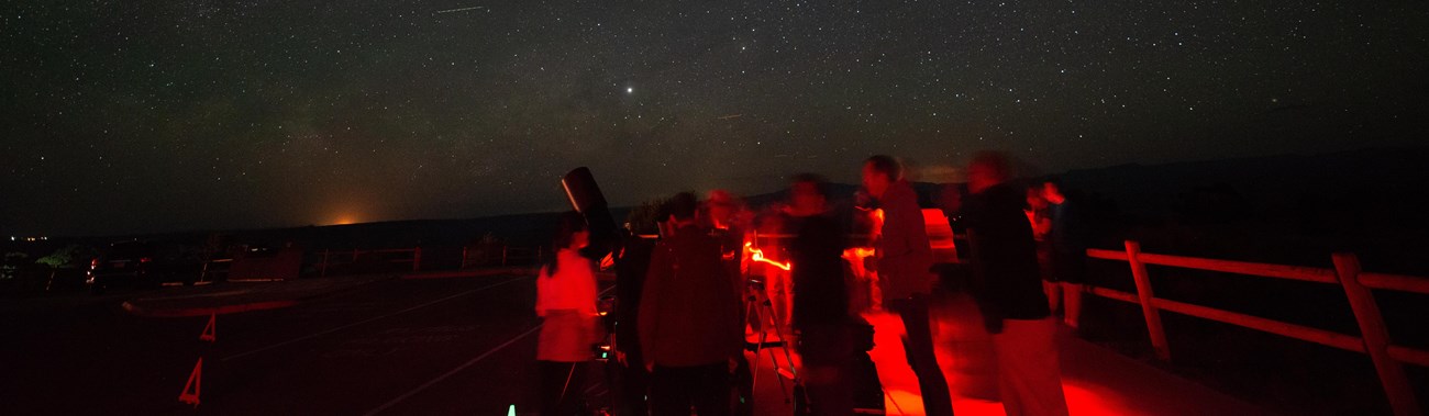 people illuminated in red gather around a telescope pointed at a starry sky