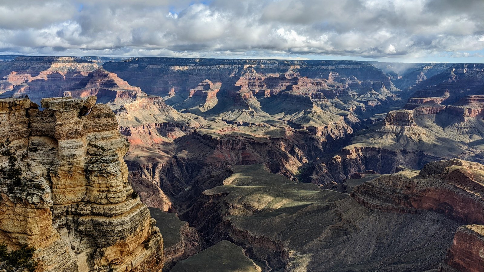 A cliff of yellow rock extends into a view of Grand Canyon\'s colorful landscape with clouds overhead