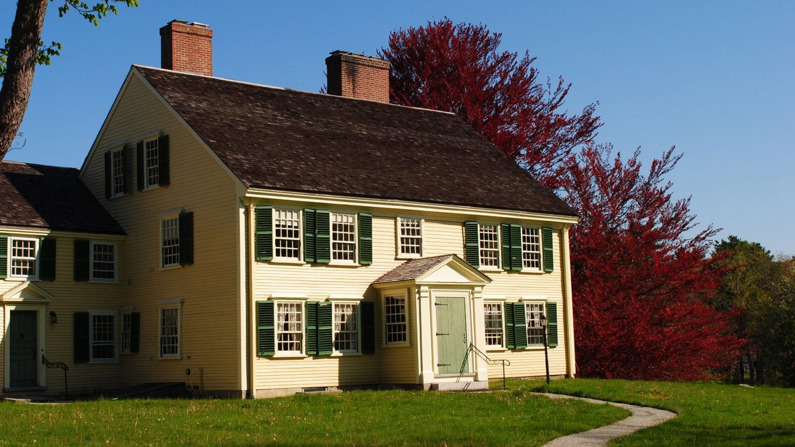 yellow house with green shutters, two stories high with a two story annex on the left. 