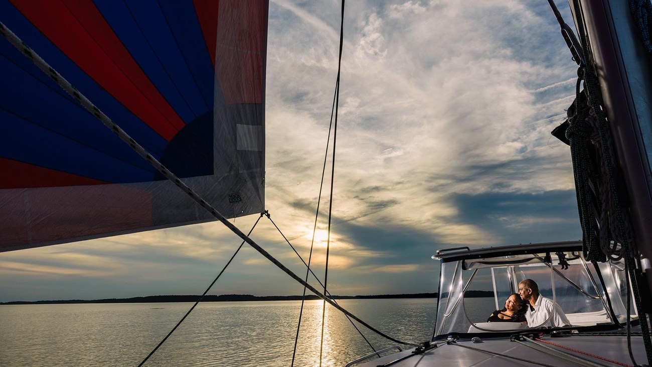 Couple on a sailboat with a colorful sail and vibrant sunset reflecting on calm waters.