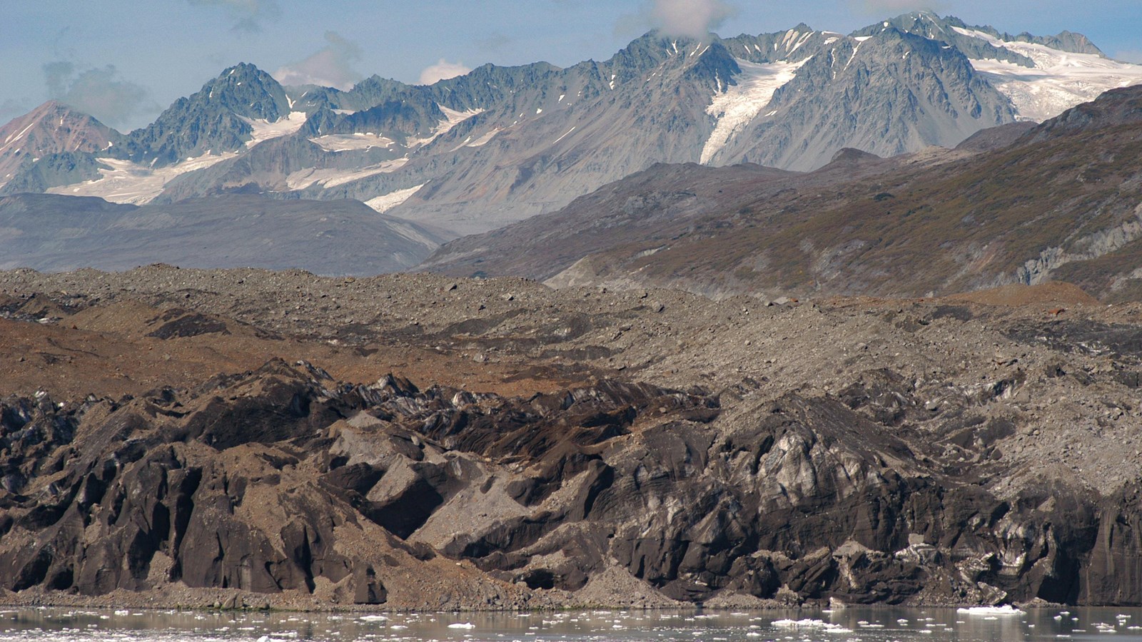 A rocky glacier is covered with brown rock. Distant mountains rise steeply in the background.