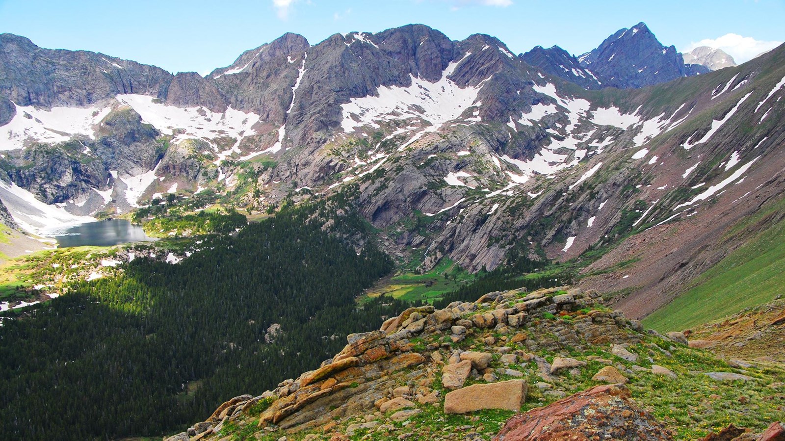 Green tundra in foreground, a forested basin with alpine lake and snowy peaks