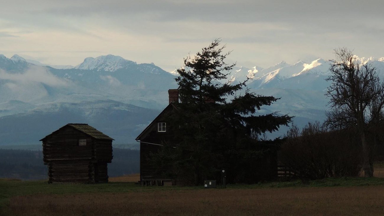 Old wood house in field with mountain range behind.
