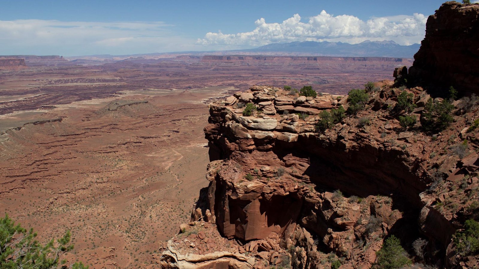 A rocky, steep cliff edge with vegetation in the foreground, far off canyons, mountains in the back.