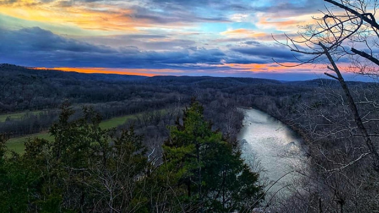 A dusk photo overlooking a river an forest with purple and pink and blue clouds as the sun sets