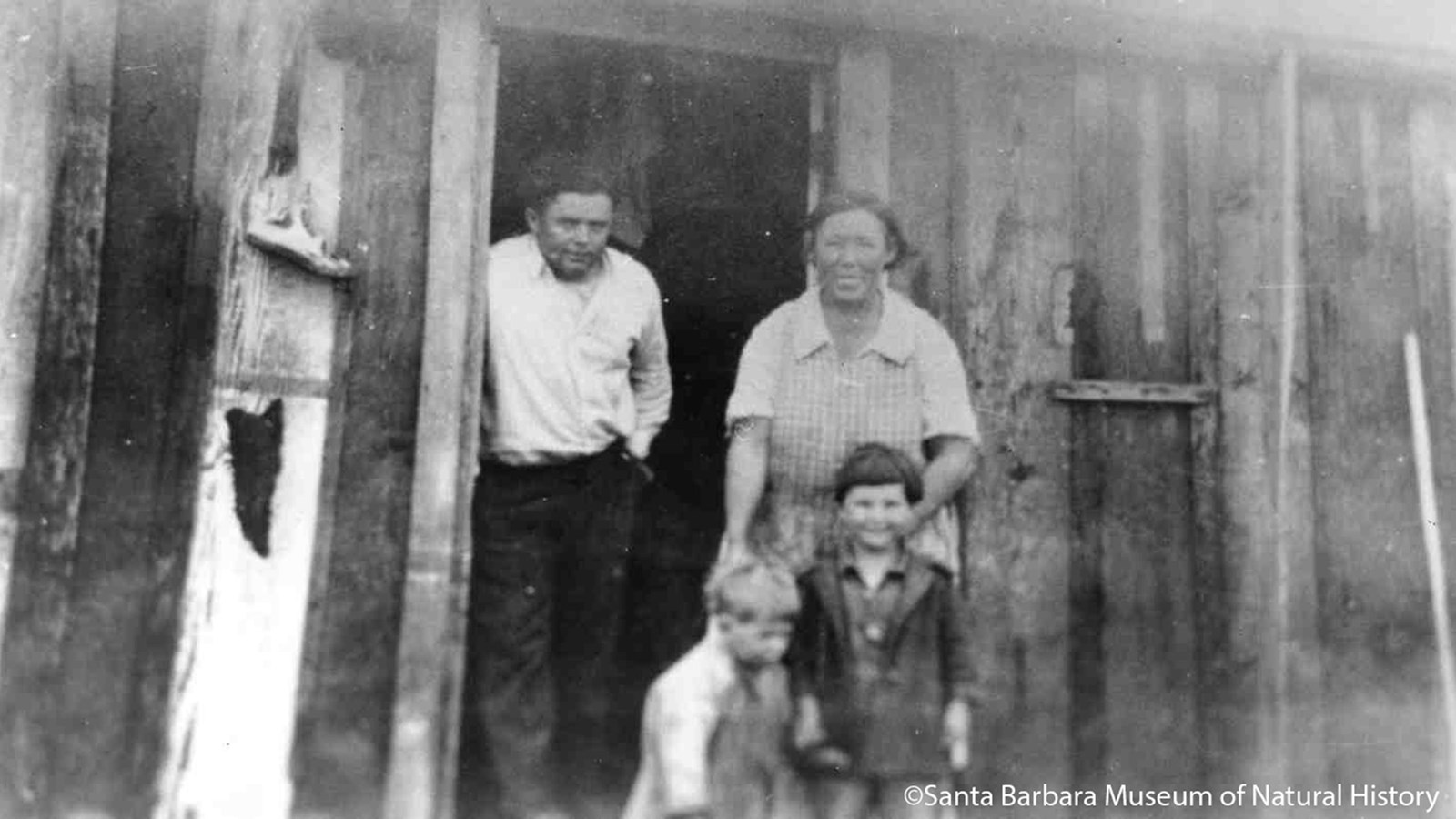 Historic photo of family standing at door of small wood building.