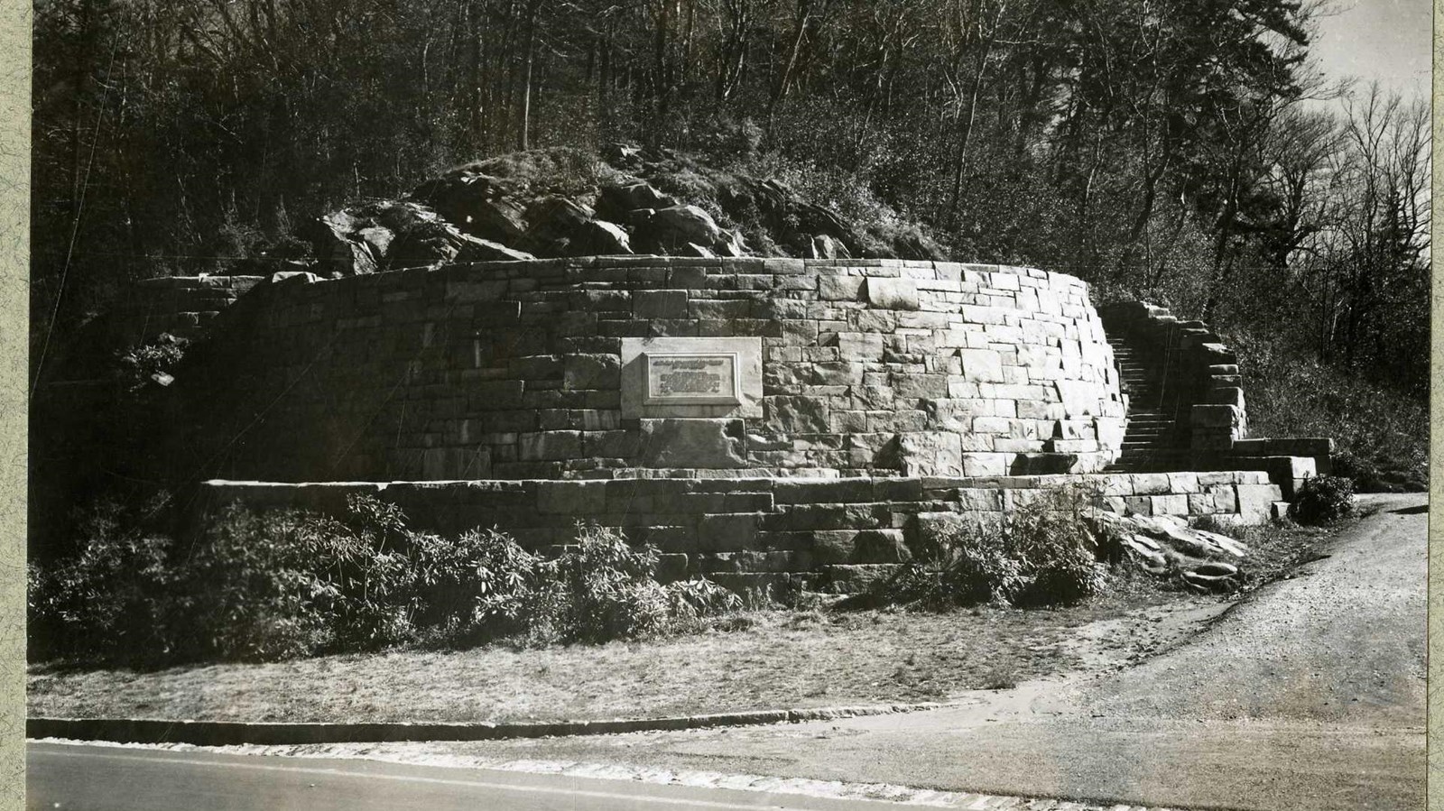 Black and white of stone memorial with plaque in middle with trees in the background