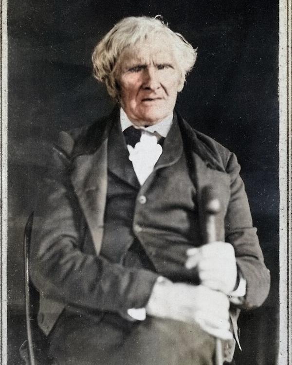 An old man with a bowl-style haircut. He sits in a nicely trimmed suit, wears a tie and holds a cane