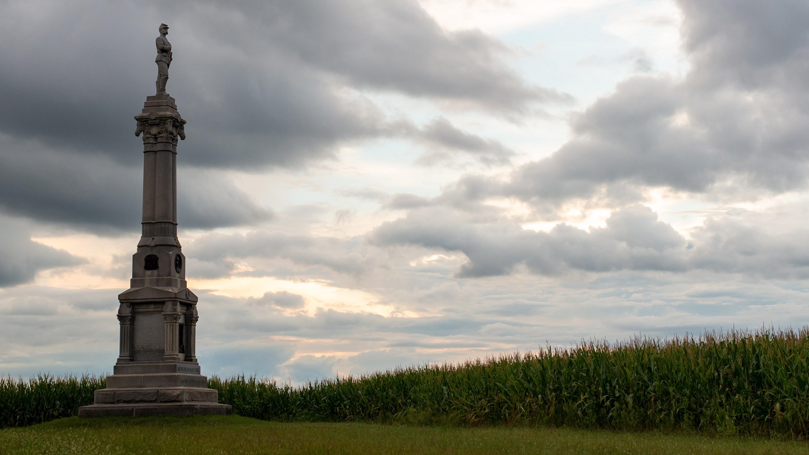A large marble statue with a soldier at the top overlooking a grass field.