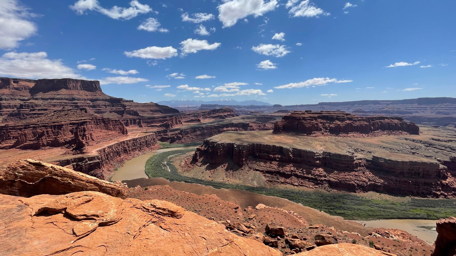 A narrow green river bends around a deep canyon on a sunny day.