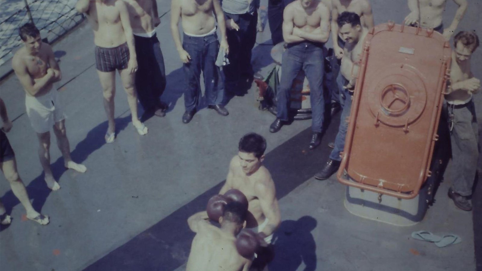 About a dozen shirtless sailors watch as two shirtless men box. A red hatch in the deck is open.