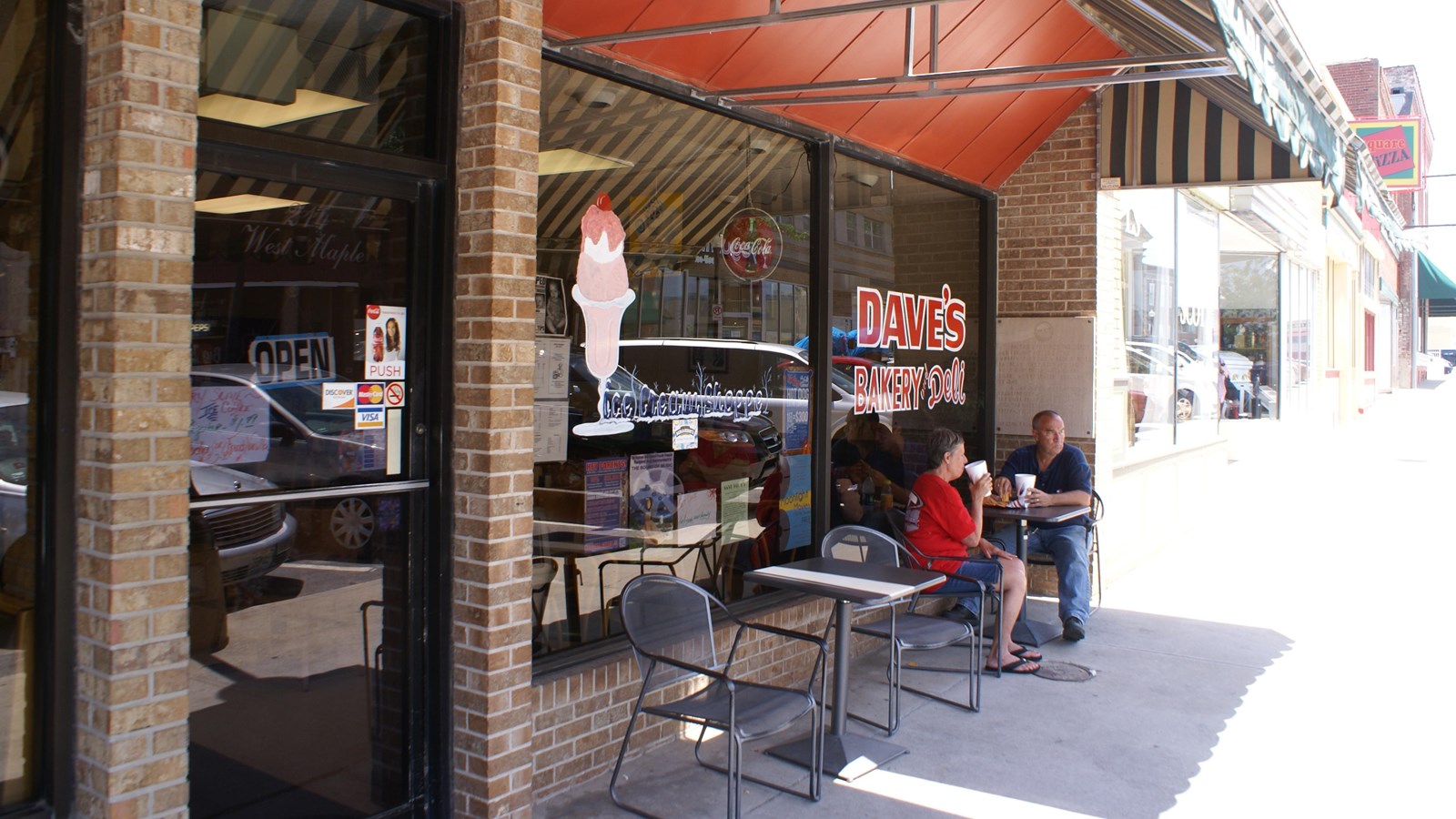 An awning hangs over an open cafe, with people sitting at small table.s