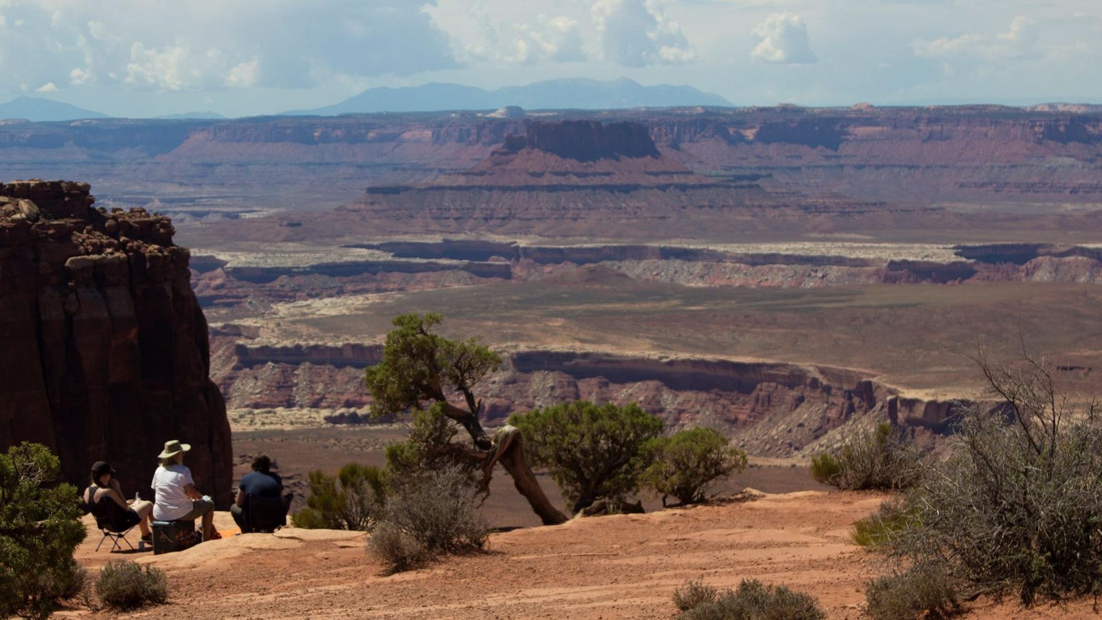 Three people sit along the edge of the overlook, sweeping canyons and clouds build in the distance