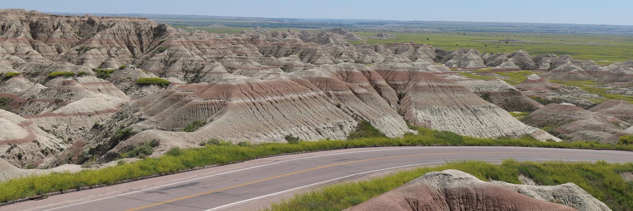 a paved road descends into a prairie landscape with buttes in the background.