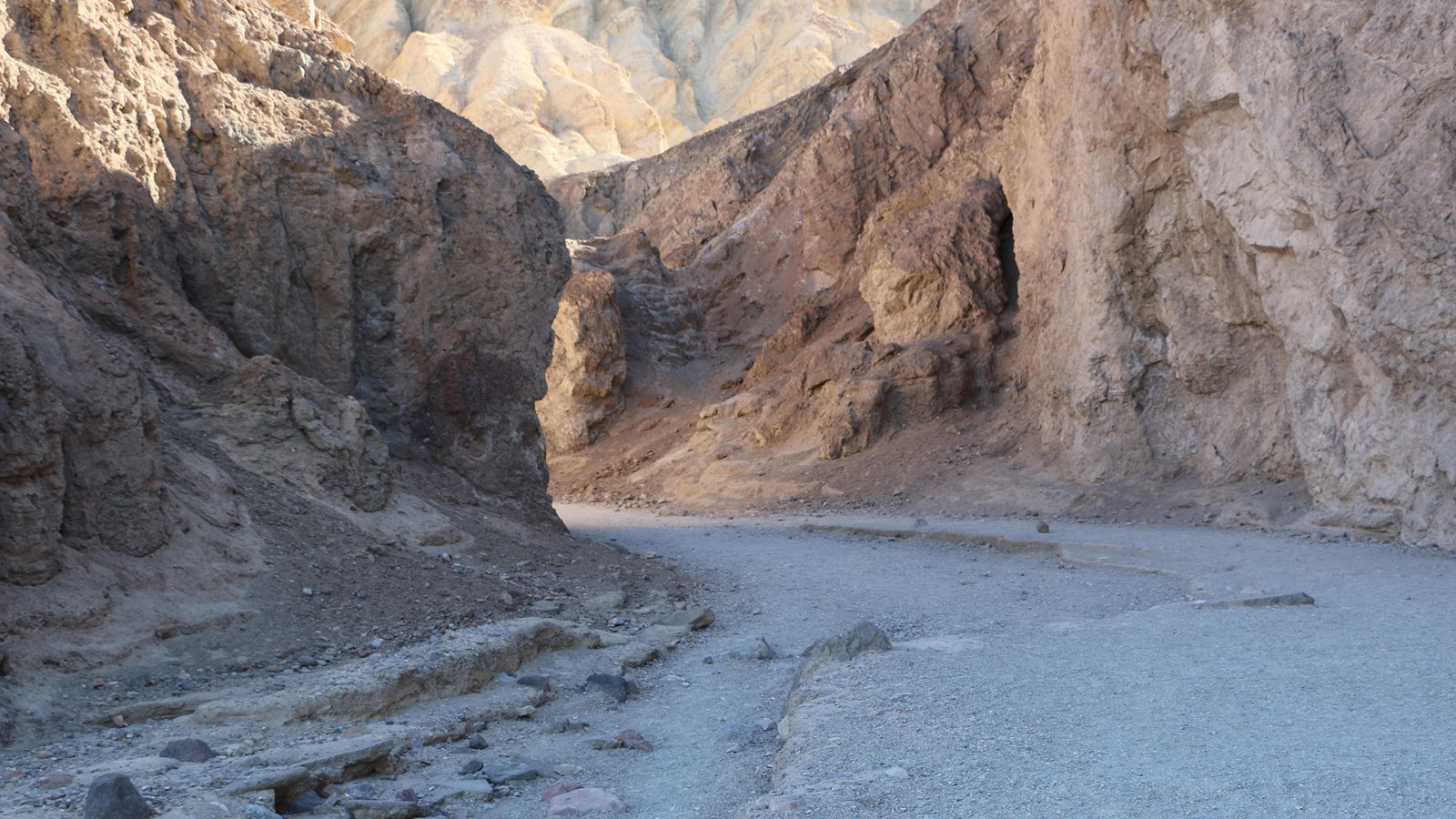 Tan canyon walls tower above a gravel wash which curves to the left deeper into the canyon.