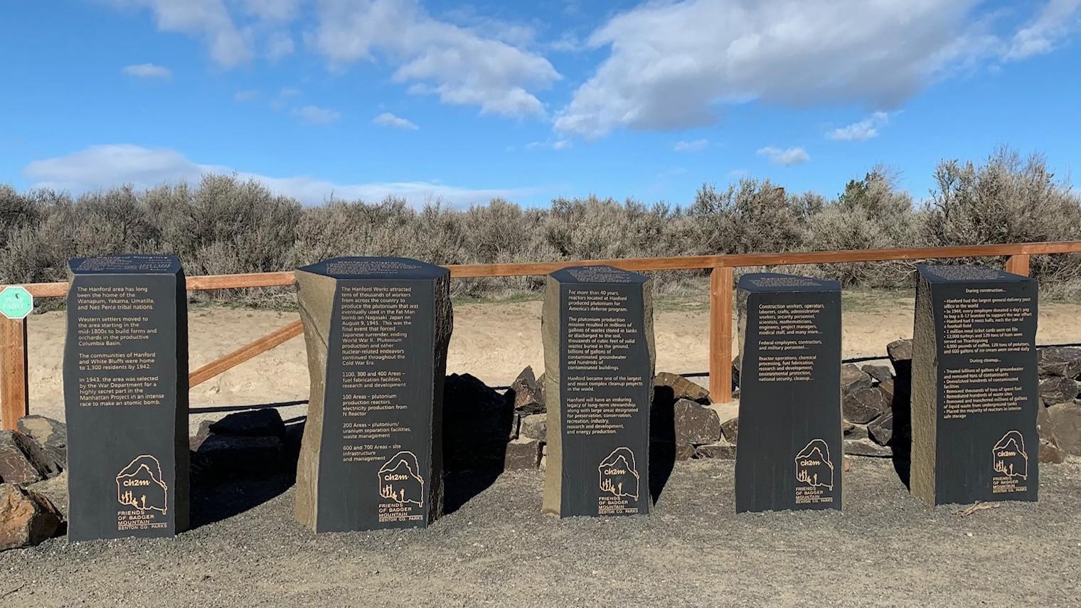 Color photograph of 4 grey stone inscribed monoliths in front of a wooden gate in a shrub steppe 
