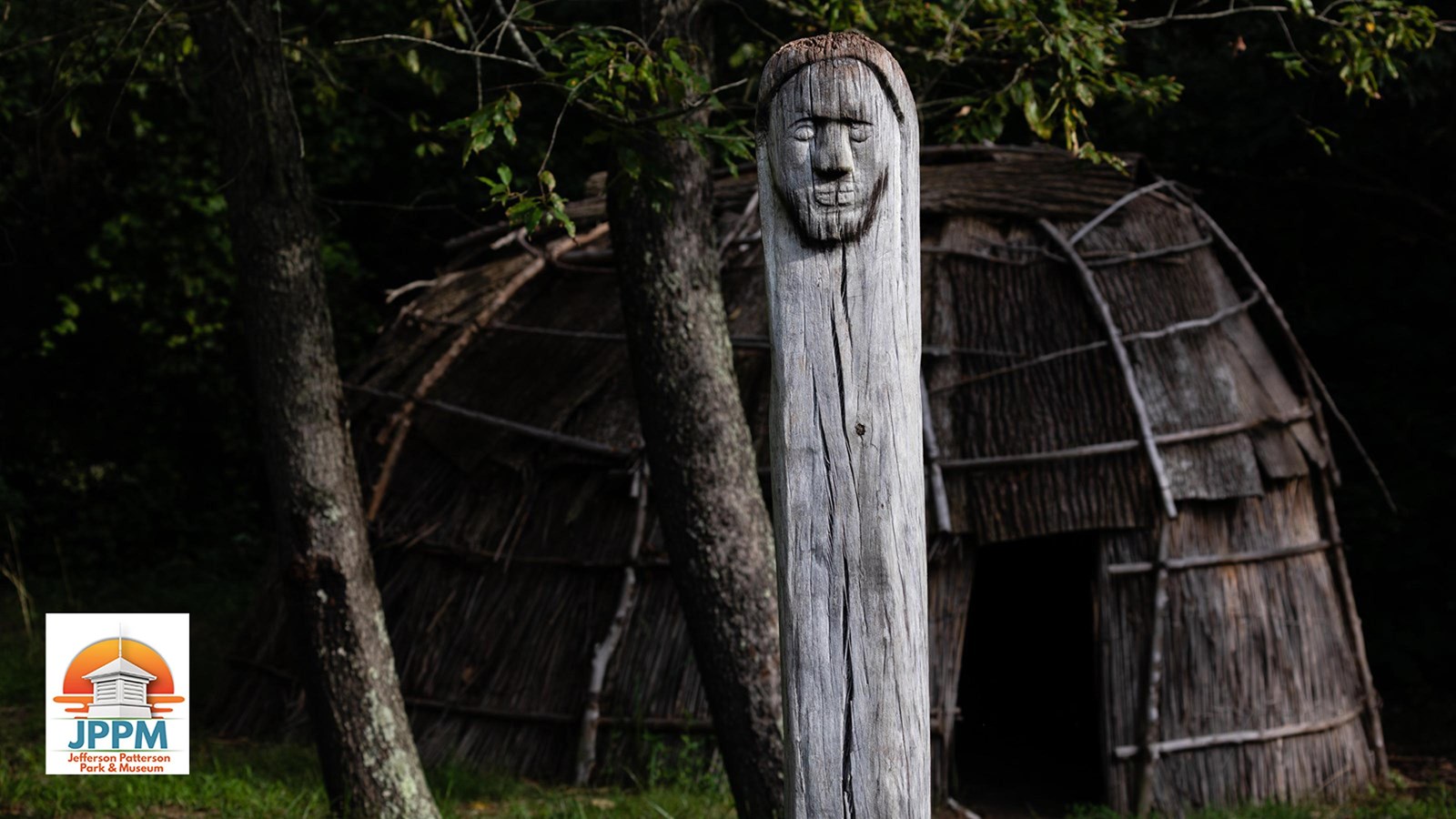 A carved wooden post with a face stands in front of a traditional thatch and bark hut.