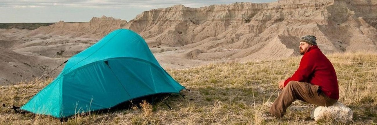 a hiker in a red jacket sits next to a blue tent with badlands buttes in the background