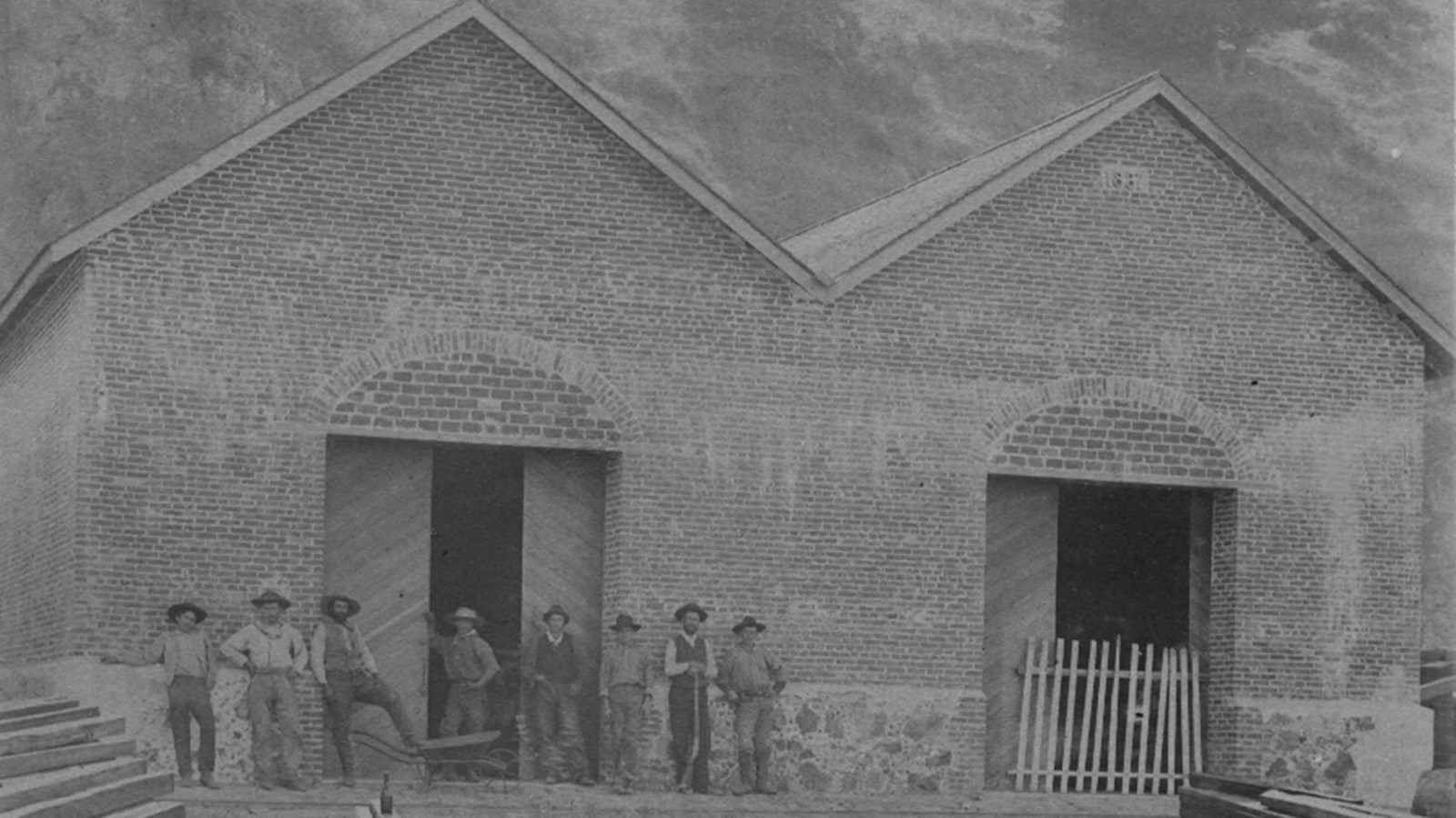 Historic photos of men posing for picture next to double gabled brick building. 