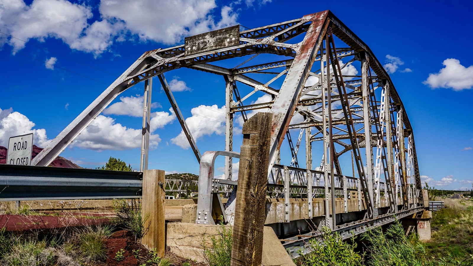 A 100 foot long steel truss bridge. In the background and red rocks, blue sky, and green shrubs.
