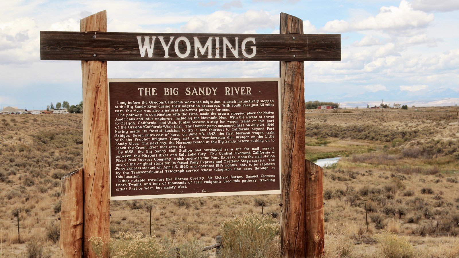 Large historic sign overlooks Big Sandy River; sagebrush, gravel, and highway visible nearby.