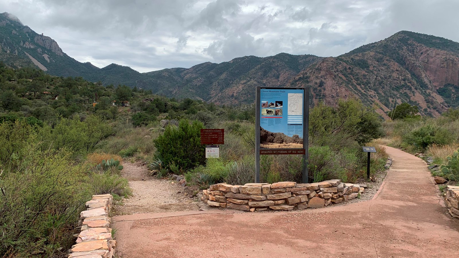 a sidewalk with stonework walls leads toward a metal trailhead sign with mountains in the background