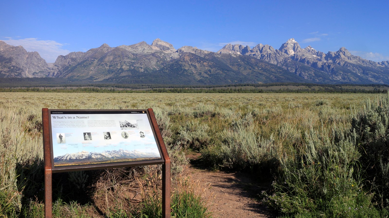 Wayside exhibit in the foreground with green sagebrush plains beyond and mountains rising beyond.