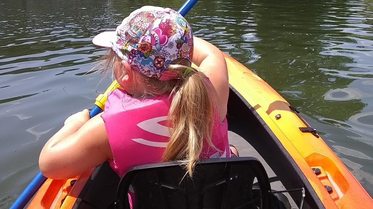 You are seated behind a little girl with a ponytail. You are in a kayak and she is holding a paddle.