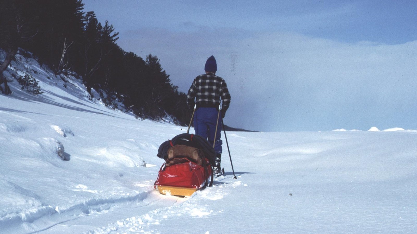 Man pulling camping equipment on sled along snowy shoreline.