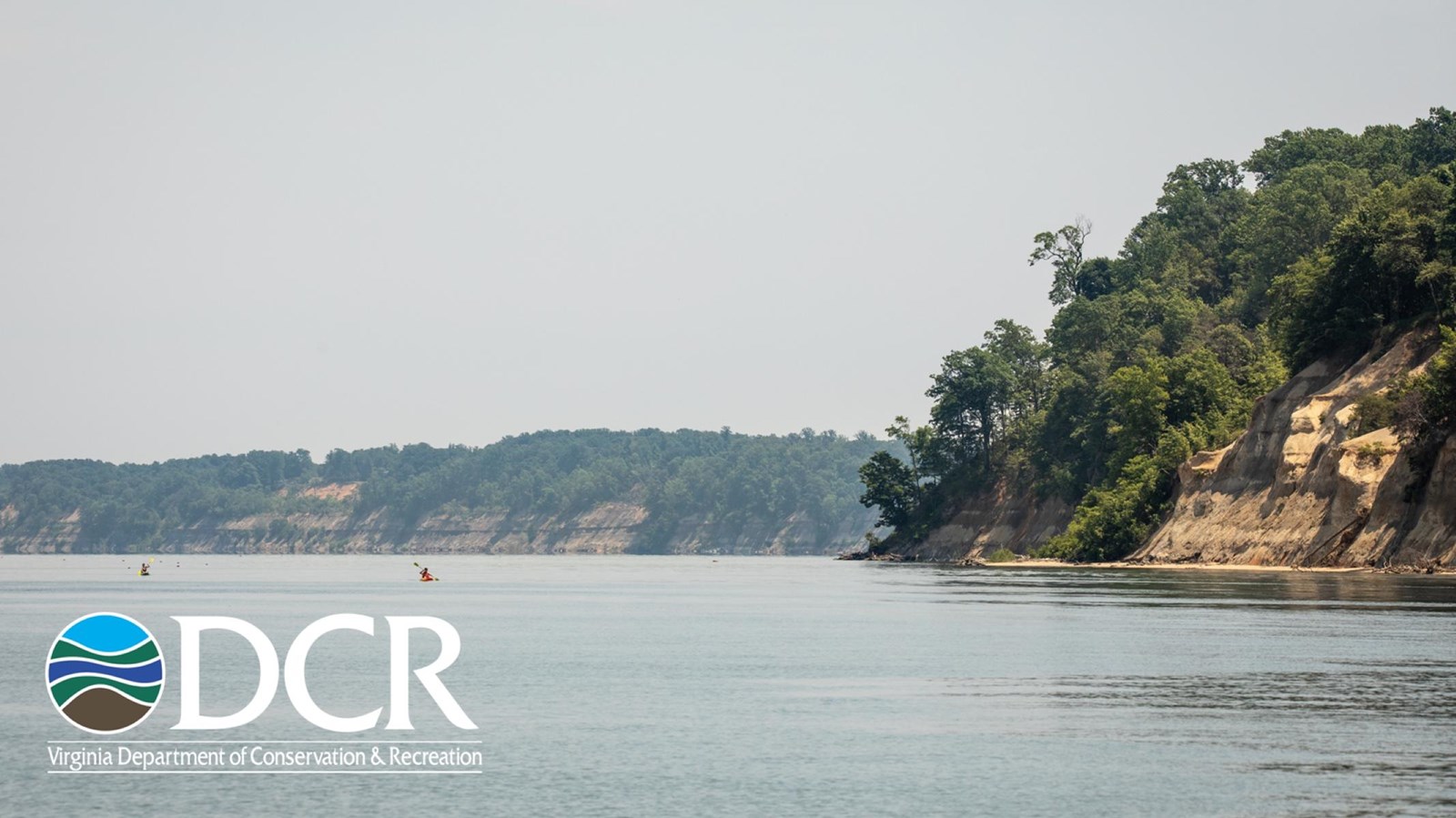 A tranquil river scene with steep, green cliffs on one side and kayakers paddling in the water.