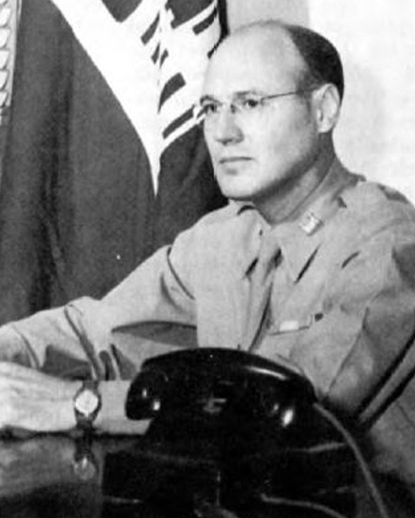 A man in uniform sits at a desk with a telephone in the foreground.