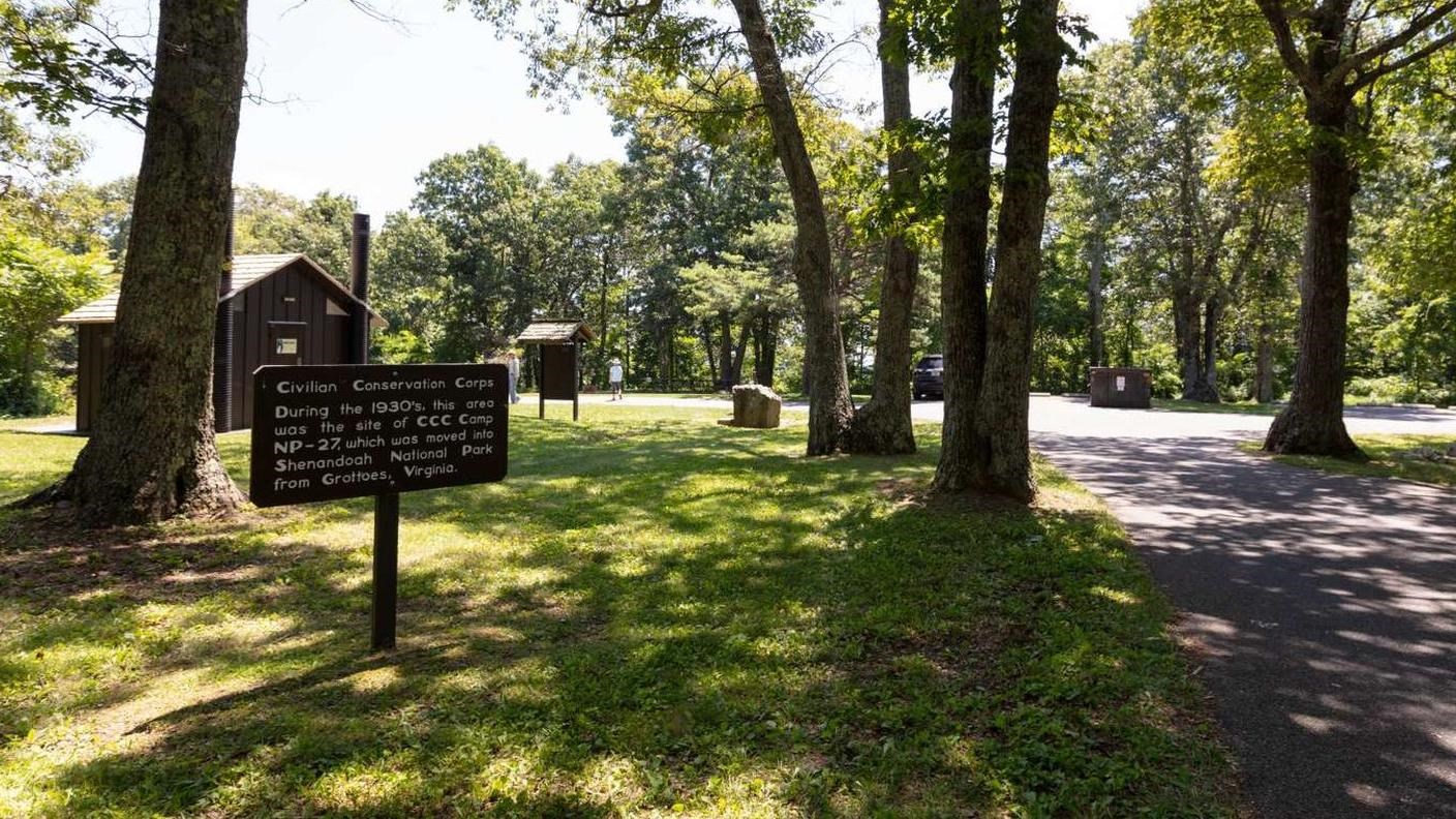 A photograph of an open green space with a brown sign, water fountain, and brown building.