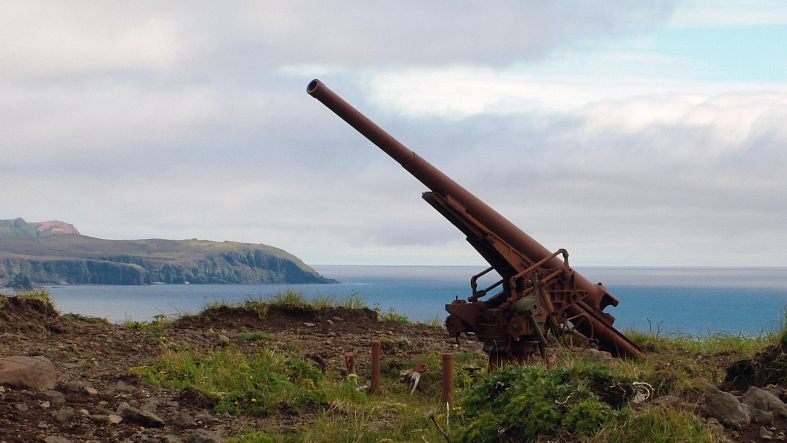 A large rusty 75 mm gun on the tundra with the ocean on the horizon.