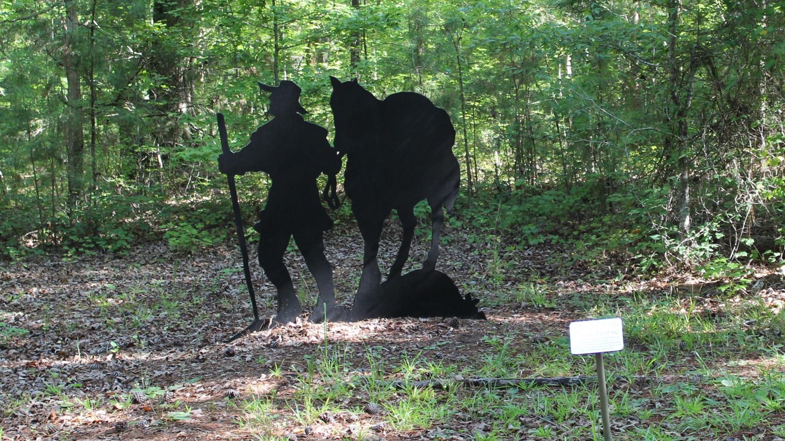 A metal cutout of a man holding a pole and pulling a mule is in the forest