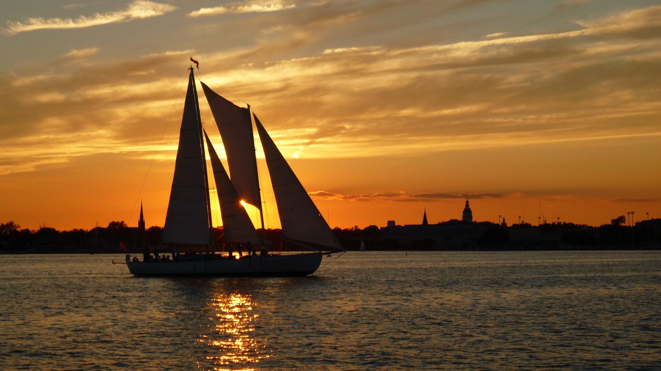 Sailboat silhouetted against a sunset with the Annapolis city skyline in the background.