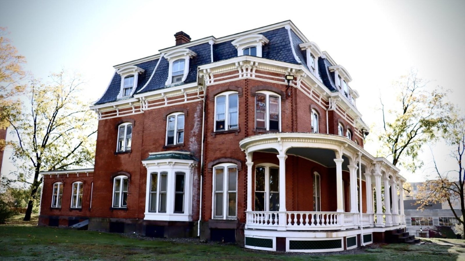 A red brick building with a covered porch and a rear ell on a sunny day during late fall. 