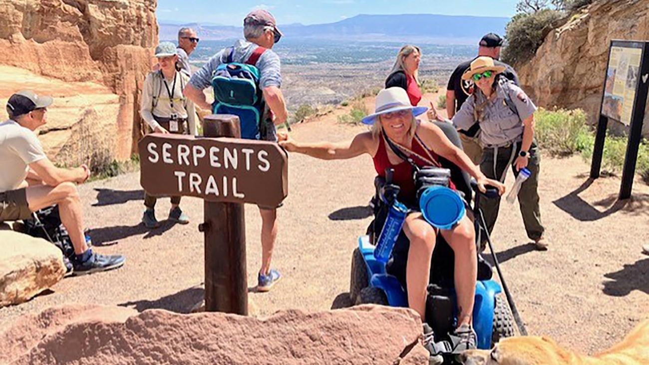 woman using wheelchair and service animal points to Serpents Trail sign with uniformed ranger behind