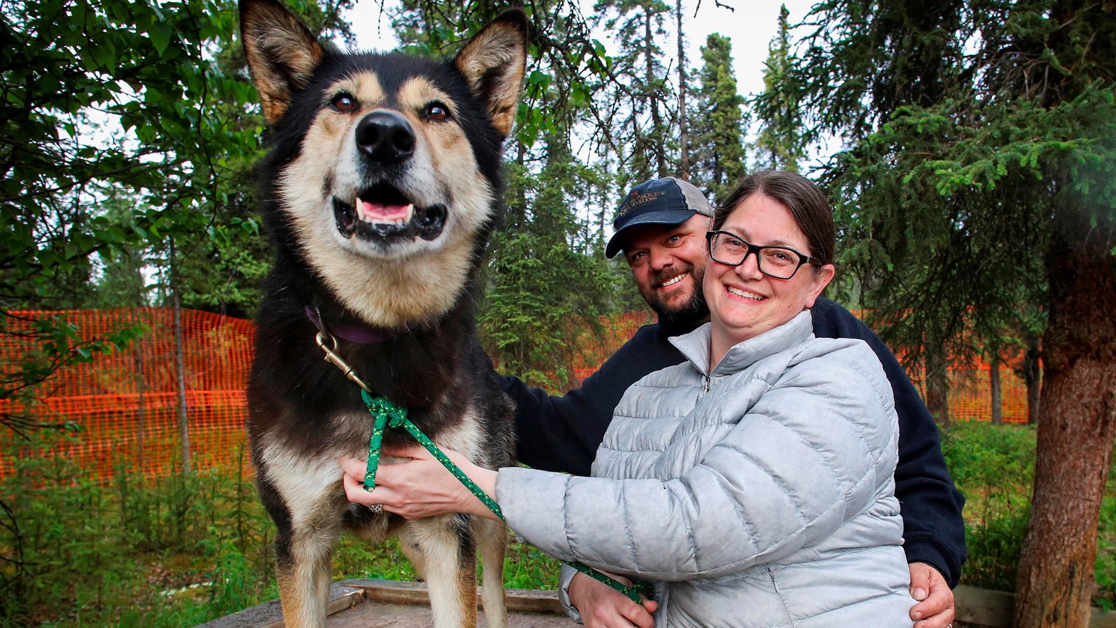 A man and a woman smile as they pet a black and brown dog. The dog is standing on a log dog house.