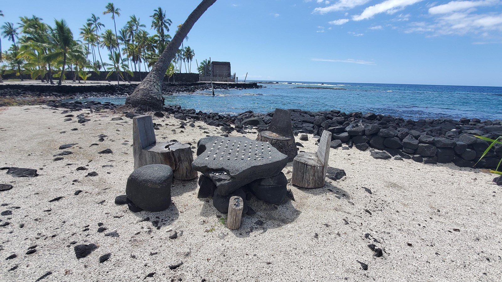 A black lava rock kōnane game board with small wooden chairs sits at the water\'s edge