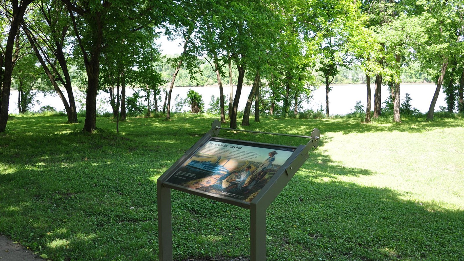Wayside on edge of sidewalk with green grass and trees on the bank of the Arkansas River.