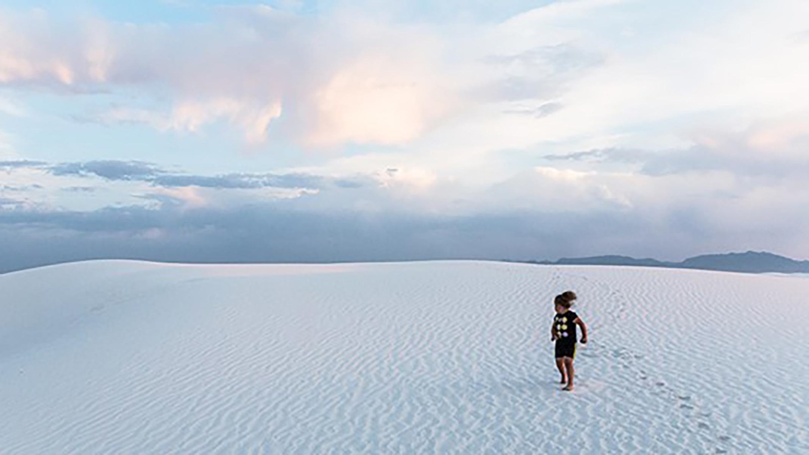 A kid leaves footprints in the sand on a partly cloudy day.