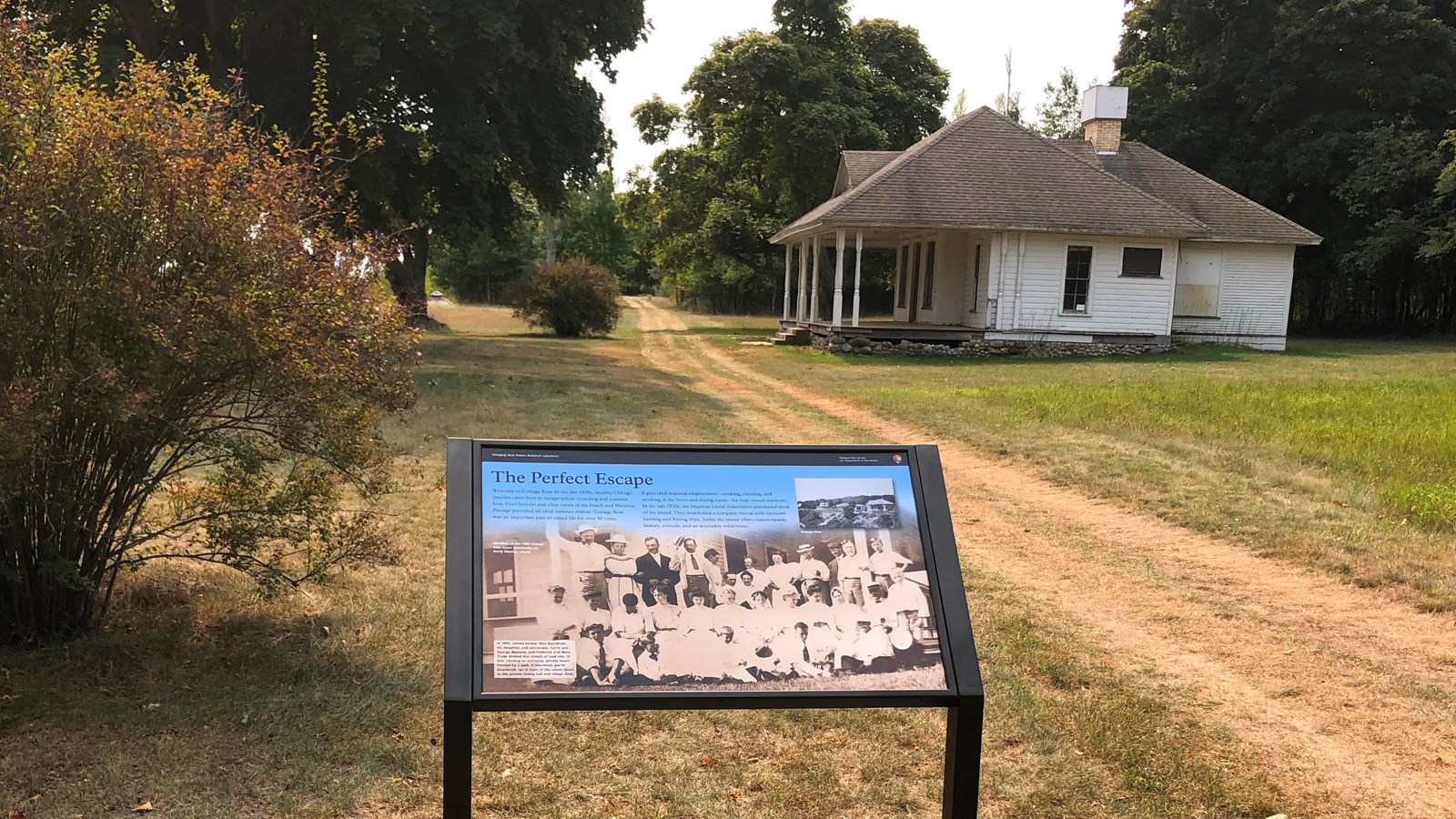 An informational sign stands alongside a wide dirt path with a historic house in the background