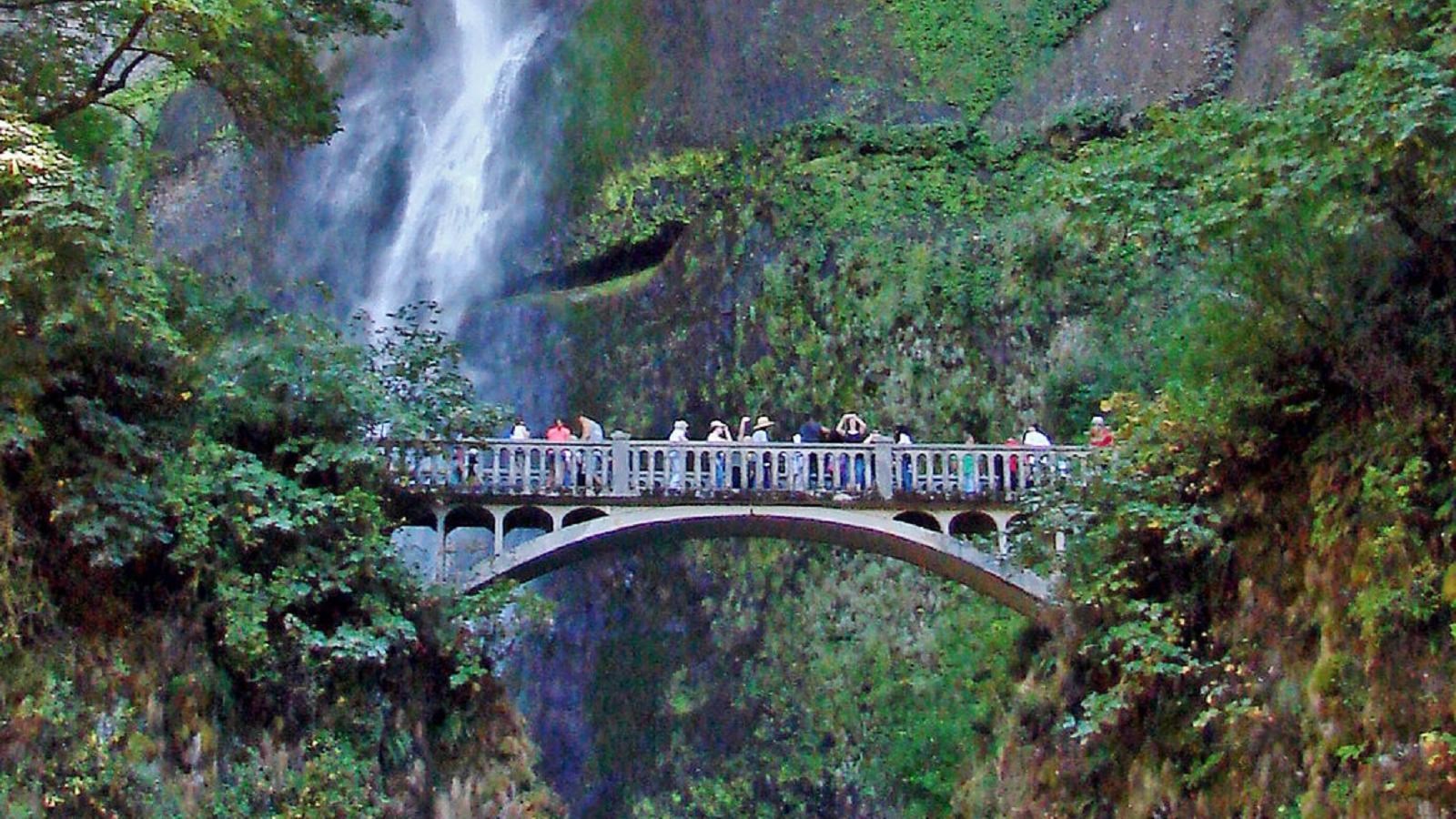A bridge spans a gap in the trail in front of a high waterfall cascading down a mountain